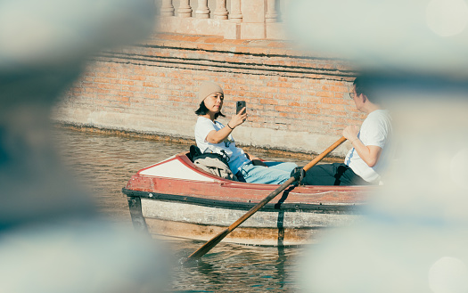 Seville, Spain; January 25th 2024: Asian tourist takes a selfie on a small boat while her husband rows. The couple enjoys a sunny day on a small boat in the Plaza de España in Seville.