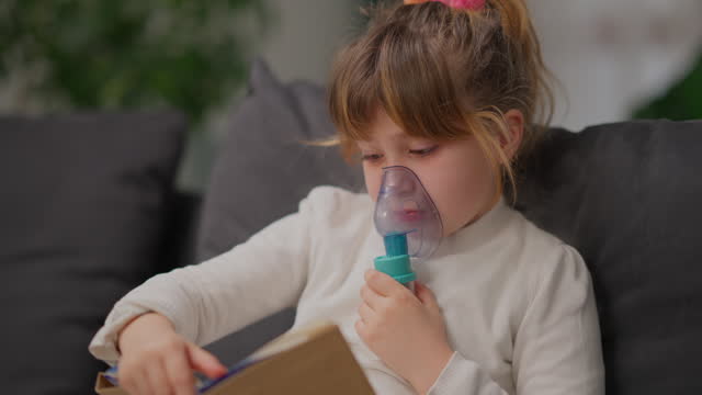 Small girl using nebulizer while enjoying reading book in the living room at home
