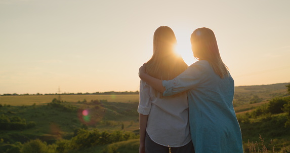 Mom hugs her teenage daughter, watching the sunset over a picturesque valley. Back view.