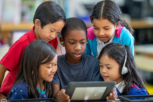 A small group of elementary students huddle around a tablet as they engage on learning online.  They are each dressed casually and focused on the screen.