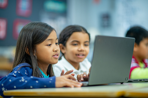 Two Elementary students are seen sitting side-by-side with a laptop open in front of them during a computer lab.  They are each dressed casually and focused on working online together.