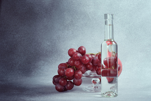 Water Bottle, Pomegranate and Red Grapes on Glass Stand, Textured Grey Background