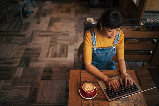 Japanese woman is sitting in a coffee bar and talking on a video call. There is coffee and a laptop on the table. High angle view