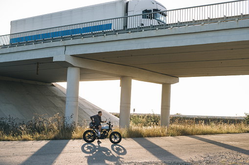Young male biker on stylish cafe racer on highway under interchange at sunset.