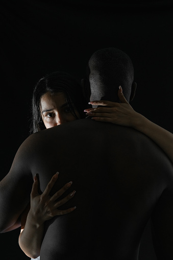 caucasian latina woman embracing african american man looking back over her shoulder, with black background