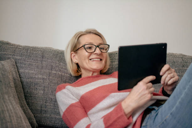 istock mature blonde woman sitting on sofa and using tablet for video call 2025879055