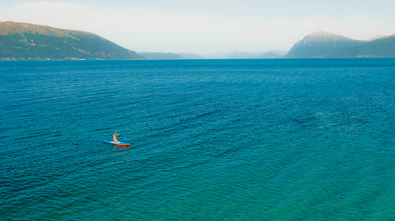 Drone high-angle photo of female on blue paddle board crossing the turquoise ocean during sunny summer day at the western fjords of Norway, Scandinavia