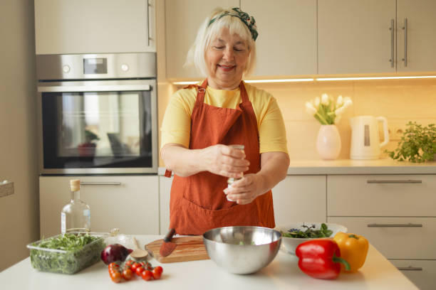 close-up of a 60s woman preparing healthy vegetable salad in kitchen, seasoning meal, adding some herbs and salt to bowl, enjoying cooking vegetarian food at home, free space for advertising text - cooking oil olive oil nutritional supplement spoon foto e immagini stock