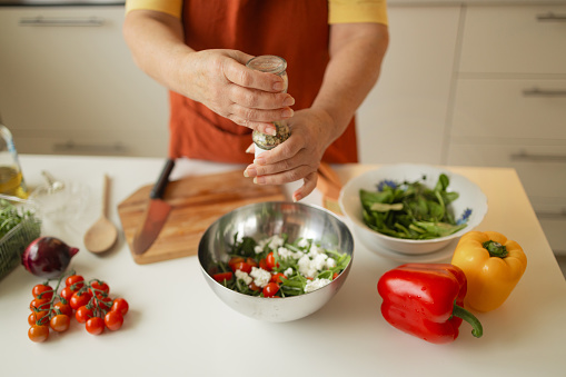 Close up of senior woman is preparing vegetable salad in the kitchen.Female adding arugula to salad. Healthy Food. Vegan Salad. Diet. Dieting Concept. Healthy Lifestyle. Cooking At Home. Prepare Food. Cutting ingredients on table