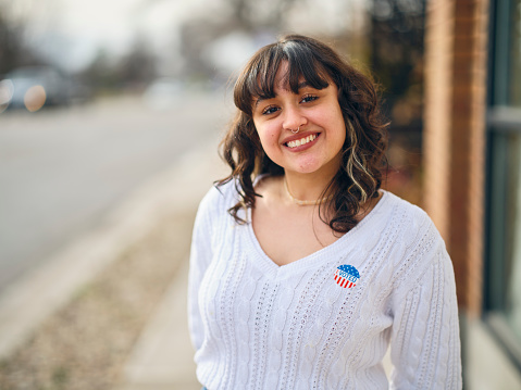A hispanic young woman standing outdoors, with an I VOTED election sticker on her sweater.