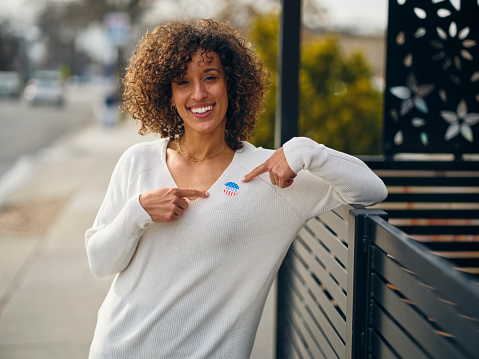 A multiracial woman standing outdoors, with an I VOTED election sticker on her sweater.