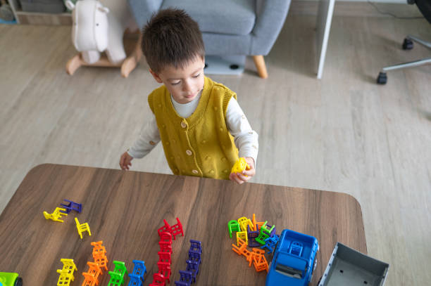 A two and half year old boy joyfully matches colorful plastic toy chairs in the comfort of his home, with a smile on his face. Engrossed in a world of vibrant hues, a beaming two-and-a-half-year-old boy gleefully orchestrates his own symphony of colors, skillfully arranging plastic toy chairs with an infectious enthusiasm, turning playtime into a delightful artistry at home.