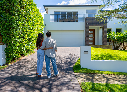 Happy Couple standing in front of their new home. They are both wearing casual clothes and embracing. Rear view. The house is contemporary with a brick facade, driveway, balcony and a green lawn. The front door is also visible.