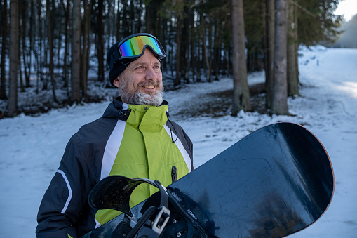Cheerful senior man with gray hair and a beard on a ski slope with snowboard in hand.