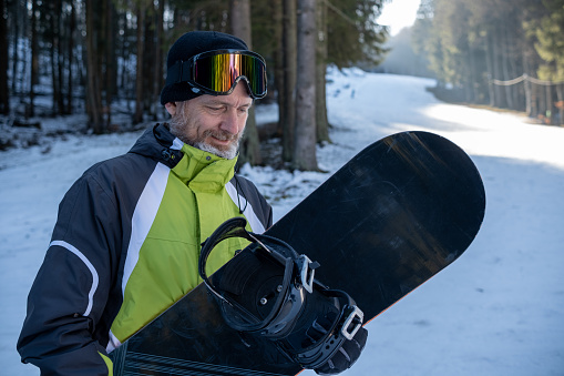 Cheerful senior man with gray hair and beard on a ski slope. He has a snowboard in his hands.