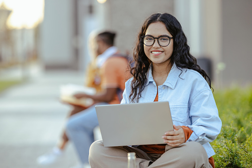 Young female student working on the laptop while sitting on the bench. Group of students is on background