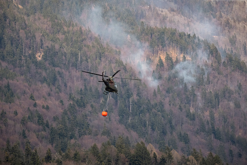 Firefighting helicopter with a bucket suspended on a cable, collecting water from a mountain lake to extinguish the flame in a forest