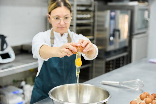 istock Young female cook in apron breaking egg into bowl while preparing dough in kitchen of bakery and cake shop 2026292565