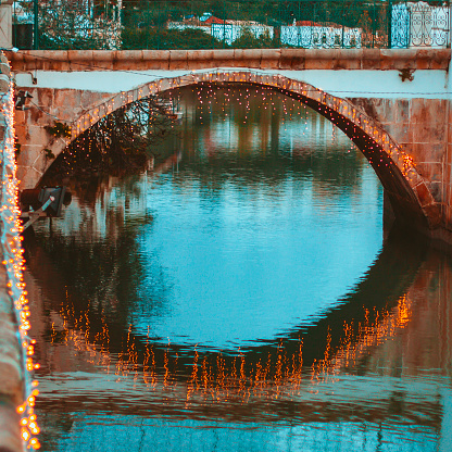 Square crop of a single span of the Roman Bridge in Tavira at twilight with decorative lights around the arches of the bridge. The lights on the bridge arch are reflected on the surface of the gently flowing Gilao river with only a few ripples on the surface.