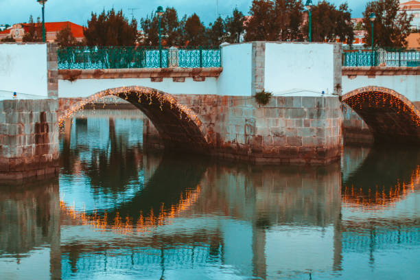 The Roman Bridge in Tavira at dusk with decorative lights around the arches of the bridge. The Gilao river is flowing gently with only a few ripples on the surface.