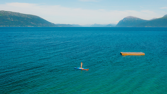 Drone high-angle photo of female on blue paddle board crossing the turquoise ocean during sunny summer day at the western fjords of Norway, Scandinavia