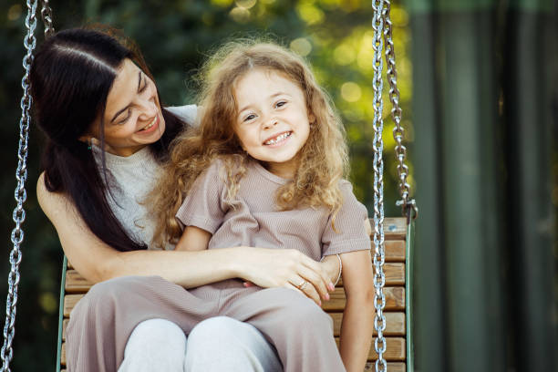 die junge mutter und ihre tochter schaukeln auf einer schaukel. kaukasische frau und kleines mädchen haben spaß auf dem spielplatz. - netting women brown hair outdoors stock-fotos und bilder