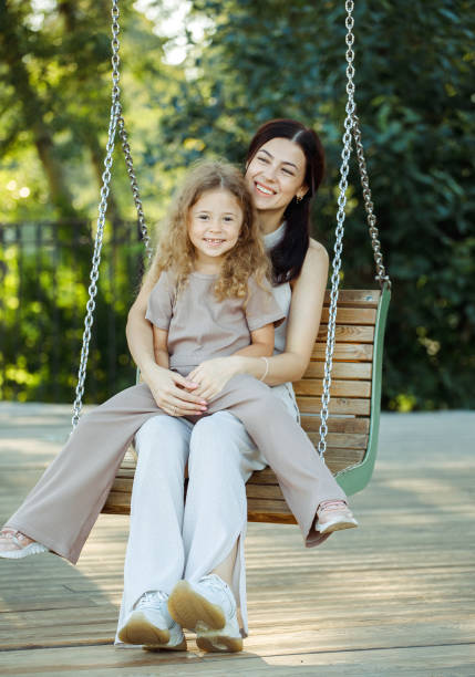 die junge mutter und ihre tochter schaukeln auf einer schaukel. kaukasische frau und kleines mädchen haben spaß auf dem spielplatz. - netting women brown hair outdoors stock-fotos und bilder