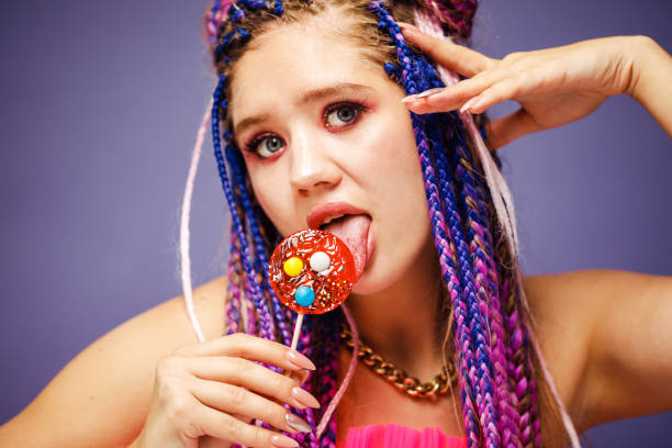 Young smiling woman with dreadlocks hairstyle and creative make-up in doll style with candy over purple background