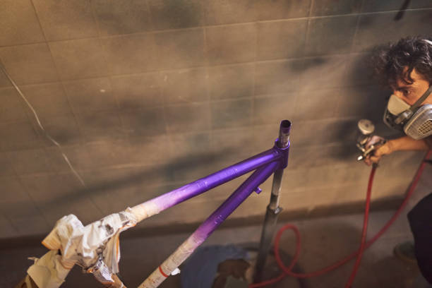 Latin young man wearing a protective respirator mask concentrated spray painting a bicycle frame in his workshop. Selective focus composition with copy space.