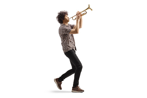 Full length profile shot of a young man walking and playing a trumpet isolated on white background