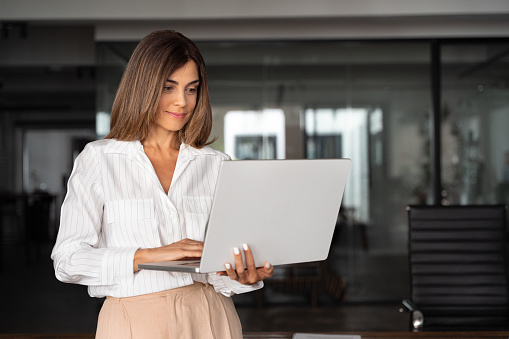 Latin Hispanic mature adult professional business woman looking at online trade app. European businesswoman CEO holding pc laptop computer using fintech tab application standing at workplace in office