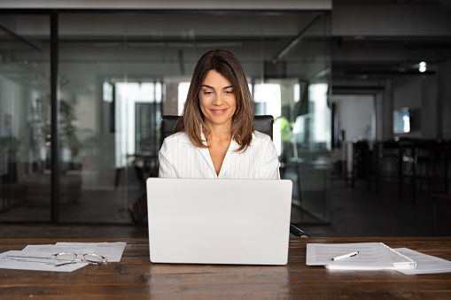 40s mid age European business woman CEO using laptop application for work sitting at table workspace in office. Smiling Latin Hispanic mature adult professional businesswoman using pc digital computer