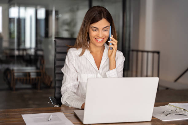 istock Smiling mature Hispanic woman sitting at table talking by mobile cellphone at workplace in office. Middle aged European Latin businesswoman having call on smartphone with business partners or clients. 2026510962