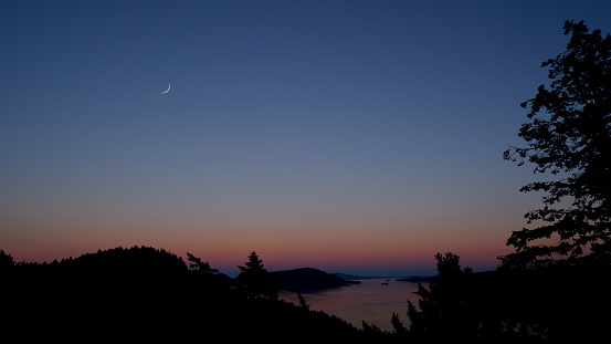 Pure blue pink and purple twilight sky spans an island bound ocean bay with a sliver of the new moon peaking out.