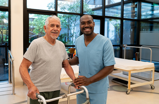 Portrait of black therapist and senior man using a mobility walker while looking at the camera smiling at a physical recovery center - Healthcare concepts