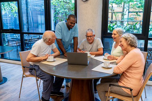 Happy group of senior friends enjoying a coffee and watching a movie on laptop while black male nurse supervises - Assisted living facilities concepts