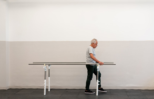 Senior man walking while leaning pn parallel bars during physical therapy - Healthcare concepts