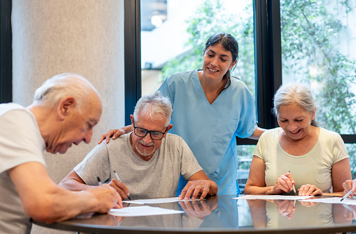 Group of happy seniors sitting around a table playing word games while cheerful nurse supervises very cheerfully - Assisted living facilities concepts