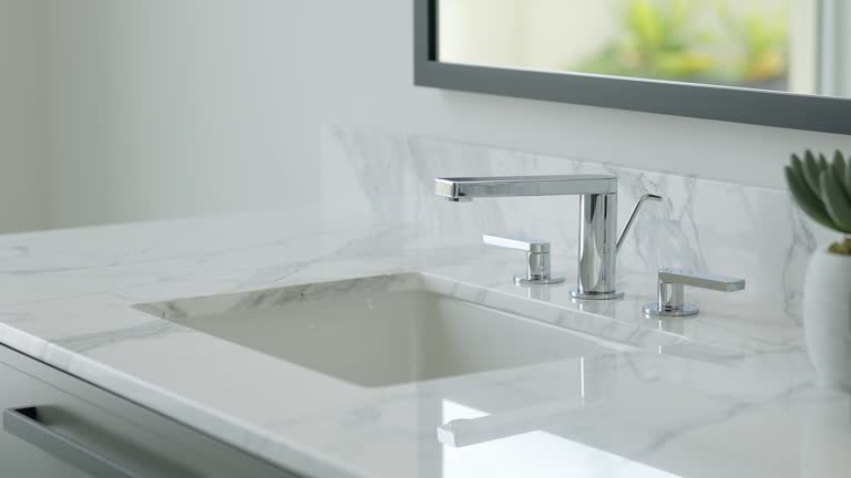 Close up of undermount ceramic washbasin and steel faucet on white marble counter with plant in pot and candle in modern luxury minimal washroom with mirror on wall