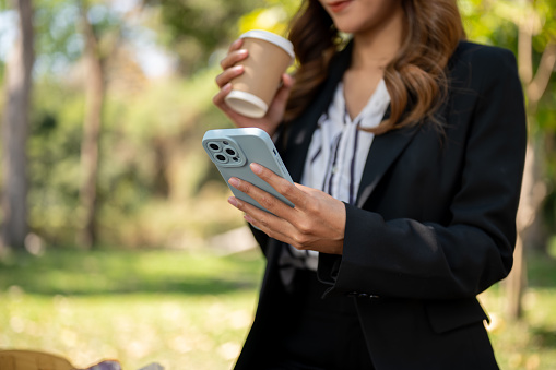 A beautiful Asian businesswoman is sipping coffee and checking messages on her smartphone while relaxing in a park. cropped shot