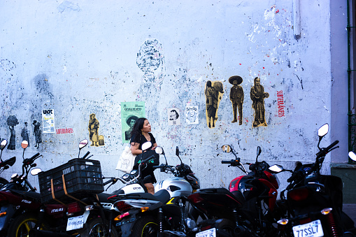 Oaxaca, Mexico: A young woman walks between a grungy wall a row of motorbikes in downtown Oaxaca.