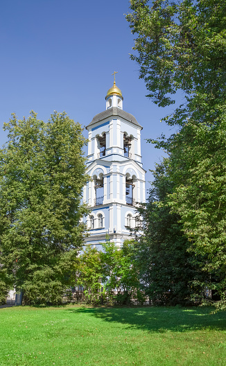 A small white bell tower surrounded by green trees against a blue sky, with a lawn in the foreground