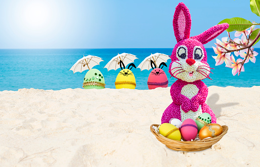 Pink easter bunny and easter egg with white umbrella on tropical beach, flower rabbit with easter egg in basket on sandy beach