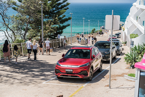 Sydney, NSW, Australia, February 17th 2024. A bustling street scene by the ocean with people walking on the sidewalk featuring a red BYD Atto 3.