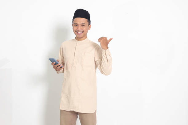 Portrait of attractive Asian muslim man in koko shirt with skullcap holding mobile phone, pointing and showing product with finger. Social media concept. Isolated image on white background stock photo