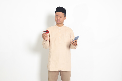 Portrait of upset Asian muslim man in koko shirt with skullcap holding a mobile phone and presenting credit card, deceived by online loans. Isolated image on white background