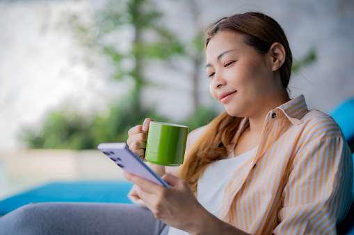 Young Asian beautiful woman sitting on sofa bed in garden, using smartphone and a cup of coffee with relaxation with atmosphere and smile of happiness at home garden, relax activity at home.