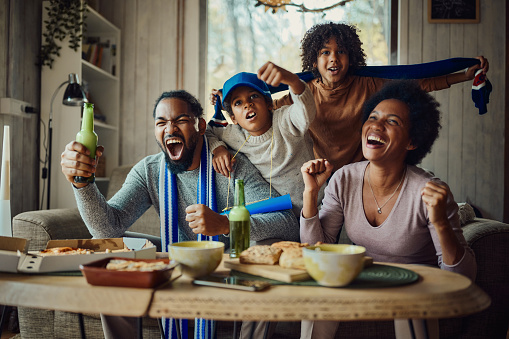 Cheerful African American family celebrating success of their favorite team while watching a game on TV at home.