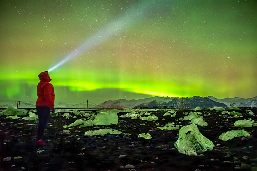 A person gazes in awe at the aurora borealis at Diamond beach, Iceland. He wears a headlamp on his head and a red jacket.