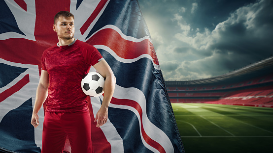 Young concentrated man, soccer payer in red uniform standing on filed with ball, representing team of England. Concept of live sport event, championship, match and tournament